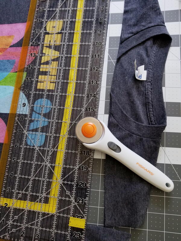 Image shows a shirt with the top cut and pulled away from the rest. It is on a mat with a ruler and rotary cutter laid on top of the shirt. Image shows a shirt with the top cut and pulled away from the rest. It is on a mat with a ruler and rotary cutter laid on top of the shirt.